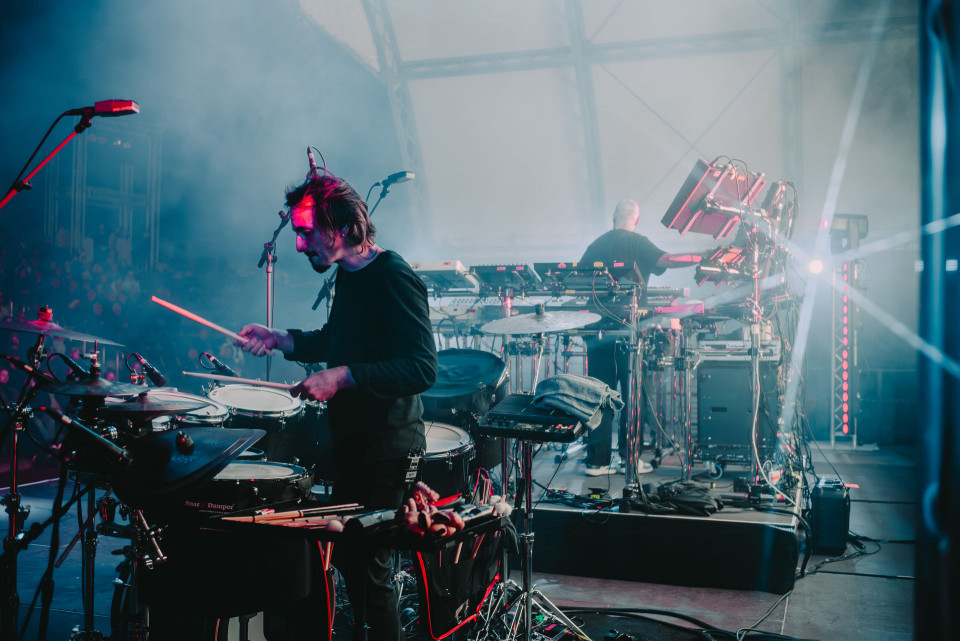Ein Schlagzeuger spielt auf einer neblig beleuchteten Konzertbühne, umgeben von Mikrofonen, Kabeln und Percussion-Instrumenten. Im Hintergrund bedient ein weiterer Musiker elektronische Geräte und Synthesizer, während farbige Scheinwerfer durch den Bühnenrauch strahlen.