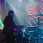 Ein Gitarrist steht im Halbdunkel auf einer Konzertbühne, während hinter ihm ein Musiker an elektronischen Geräten spielt. Bunte Lichtstrahlen, Nebel und großflächige pink-blaue Projektionen erzeugen eine atmosphärische Live-Show.