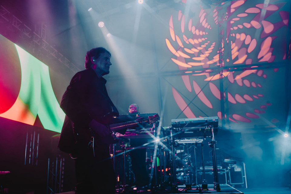 Ein Gitarrist steht im Halbdunkel auf einer Konzertbühne, während hinter ihm ein Musiker an elektronischen Geräten spielt. Bunte Lichtstrahlen, Nebel und großflächige pink-blaue Projektionen erzeugen eine atmosphärische Live-Show.