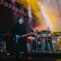 Ein Musiker spielt auf einer Bühne E-Gitarre, während im Hintergrund ein weiterer Künstler an mehreren Keyboards und elektronischen Geräten steht. Bunte Scheinwerfer, Nebel und große Lichtprojektionen erzeugen eine dramatische Konzertatmosphäre.