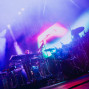 Auf einer Konzertbühne stehen ein Schlagzeug, Keyboards und Mikrofone im intensiven violett-blauen Licht. Bunte Scheinwerfer und Nebel erzeugen eine dynamische, atmosphärische Live-Performance-Stimmung.