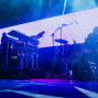 Auf einer dunkel beleuchteten Bühne spielen zwei Musiker zwischen Synthesizern und einem Schlagzeug. Im Hintergrund leuchtet eine große LED-Wand mit pink-blauen geometrischen Mustern, während blaue Scheinwerfer die Szene durchfluten.