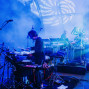 Auf einer blau beleuchteten Konzertbühne spielt ein Musiker Schlagzeug, während im Hintergrund ein weiterer Musiker an elektronischen Instrumenten steht. Nebel, Mikrofone, Kabel und starke Lichtstrahlen erzeugen eine atmosphärische Live-Performance vor Publikum.
