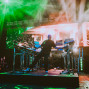 Ein Musiker steht auf einer Konzertbühne zwischen zahlreichen Synthesizern und elektronischen Geräten, während grüne Laser und Nebel die Szene beleuchten. Im Hintergrund läuft auf einer großen LED-Wand eine orangefarbene Visualisierung, und weitere Instrumente wie Gitarren und Schlagzeug sind seitlich zu sehen.