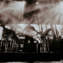Ein Musiker steht auf einer Bühne zwischen Synthesizern und elektronischem Equipment, umgeben von dichtem Nebel. Helle Scheinwerfer strahlen von oben durch den Rauch und erzeugen eine dramatische Konzertatmosphäre.