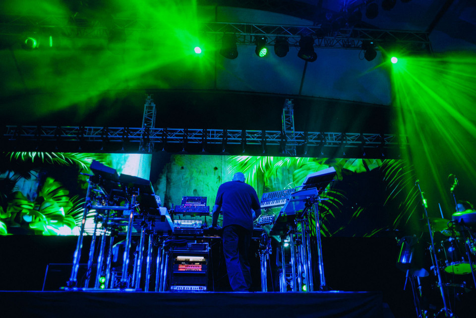 Auf einer Konzertbühne steht ein Musiker von hinten vor Synthesizern und elektronischem Equipment, während grüne Laserstrahlen und blaues Licht die Szene erhellen. Im Hintergrund sind große LED-Projektionen mit tropischen Blattmustern sowie Schlagzeug und Bühnenkonstruktionen zu sehen.