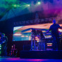 Auf einer Konzertbühne stehen zwei Musiker zwischen Synthesizern, Gitarren und technischem Equipment. Bunte Lichtstrahlen, Nebel und eine große LED-Leinwand erzeugen eine intensive, elektronische Live-Atmosphäre.