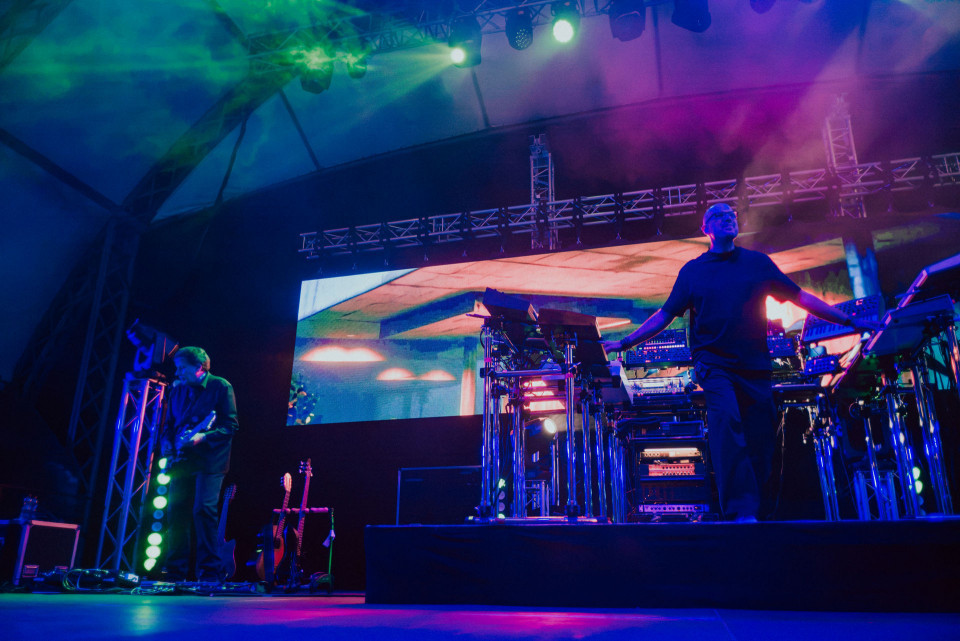 Auf einer Konzertbühne stehen zwei Musiker zwischen Synthesizern, Gitarren und technischem Equipment. Bunte Lichtstrahlen, Nebel und eine große LED-Leinwand erzeugen eine intensive, elektronische Live-Atmosphäre.
