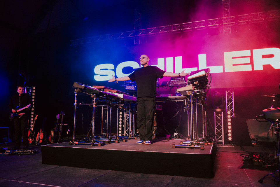 Ein Musiker steht mit ausgebreiteten Armen zwischen mehreren Keyboards und elektronischen Geräten auf einer bunt beleuchteten Bühne. Im Hintergrund leuchtet groß der Schriftzug „SCHILLER“, während Nebel und rote sowie blaue Lichter die Konzertatmosphäre prägen.