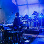 Auf einer blau beleuchteten Bühne stehen Musiker zwischen Schlagzeug, Mikrofonen, Synthesizern und vielen Kabeln. Links ist ein großes Publikum zu sehen, während Nebel und Scheinwerfer die Konzertatmosphäre verstärken.