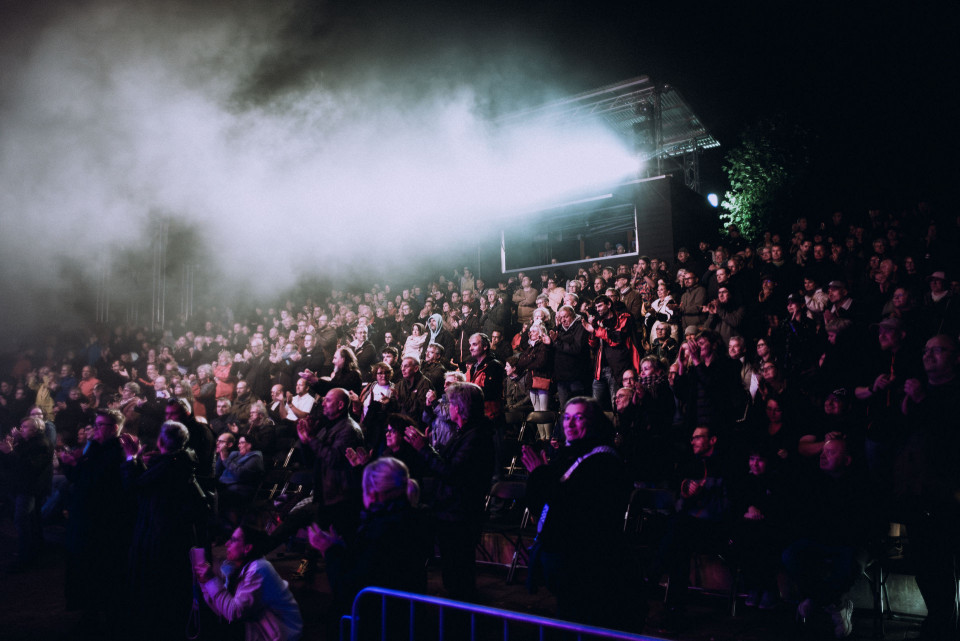 Eine große Menschenmenge sitzt und steht bei einer nächtlichen Open-Air-Veranstaltung und applaudiert. Über dem Publikum liegen dichter Nebel und grelle Scheinwerferstrahlen, die die Szene dramatisch beleuchten.