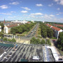Das Foto zeigt eine weite Stadtansicht von einem Dach aus, mit einer breiten, von Bäumen gesäumten Straße und mehreren Wohn- und Geschäftsgebäuden. Am Himmel sind viele kleine Wolken zu sehen, während unten Autos auf den Straßen fahren.