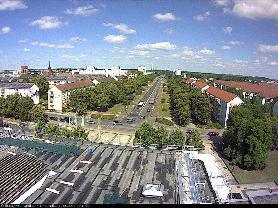 Das Foto zeigt eine weite Stadtansicht von einem Dach aus, mit einer breiten, von Bäumen gesäumten Straße und mehreren Wohn- und Geschäftsgebäuden. Am Himmel sind viele kleine Wolken zu sehen, während unten Autos auf den Straßen fahren.