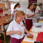 Auf einem Markttstand schneiden mehrere Personen Brot und Kuchen in Anwesenheit eines Holzschildes mit dem Logo „Bäcker Schreiber“. Vor dem Stand sind viele verschiedene Brotsorten und Kuchen aufgereiht.