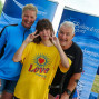 Drei Personen – eine Frau und zwei Kinder – posieren vor einem blauen Banner mit dem Logo einer Behindertensportorganisation. Die gelbe T-Shirt der Kind trägt ein „Love“-Motiv.