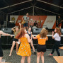 Auf einer Bühne, die mit einem Zeltdach bedeckt ist, spielen eine Band mit mehreren Musikern. Vor der Bühne tanzen eine Gruppe junger Leute ausgelassen und mit viel Freude.