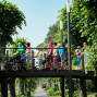 Auf einem erhöhten Holzbalkon gehen eine Gruppe von Menschen, darunter Kinder und ältere Personen, zu Fuß. Um den Balkon herum wachsen dichte, grüne Büsche und Bäume, die einen schattigen Hintergrund bilden.