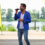 Ein Mann mit Bart trägt einen blauen Blazer, einen weißen Hemd und blaue Jeans und singt vor einem Mikrofon auf einer Veranda. Im Hintergrund ist ein See und Grünland zu sehen.