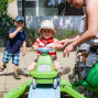 Ein kleiner Junge sitzt auf einem grünen Dreirad und wird von seiner Mutter geholfen, sich hochzuziehen. Im Hintergrund stehen ein weiterer Junge und ein Mann in einer Flanellhemd, während eine Frau mit einem Baby in einer Trage im Vordergrund steht.