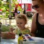 Eine Frau sitzt mit einem kleinen Kind an einem Tisch und hilft ihm, ein Bild zu malen. Im Hintergrund ist ein Outdoor-Festival mit verschiedenen Ständen und Gäste zu sehen.