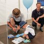 Zwei Männer und ein Junge sitzen auf dem Boden und beschäftigen sich mit einem Puzzle und einer Anleitung. Ein großer blaue Luftballon und ein Bildschirm mit Zeitangaben sind im Hintergrund zu sehen.