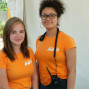 Zwei Frauen, eine mit blonden Haaren und die andere mit einem kurzen Afrikanerhaarschnitt, stehen dicht hintereinander und lächeln in die Kamera. Beide tragen orangefarbene T-Shirts mit dem Text „IN ONTA INFO“ und scheinen an einer Veranstaltung teilzunehmen.