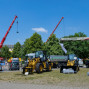 Auf dem Foto sieht man einen großen Bauzelt mit einem Kran und einem gelben Bagger, umgeben von verschiedenen Maschinen, Autos und einem Marktstand. Im Hintergrund ist ein strahlend blauer Himmel mit vereinzelten Bäumen zu erkennen.