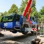 Ein großer, blauer Krans auf einem LKW steht auf einer Pflasterfläche in einer Grünanlage. Der Kran ist mit dem Firmenlogo "BKL" und weiteren Informationen versehen und neben Schutzplaketten und Leitern stehen.