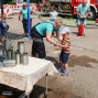 Auf dem Foto sieht man eine Frau, die einem kleinen Jungen hilft, mit einem Wasserstrahl Kanister umzuwerfen. Im Hintergrund steht ein Feuerwehrauto und es scheint sich um eine Wasserspielaktion mit mehreren Kindern zu handeln.