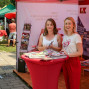 Zwei Frauen lächeln an einem roten Tisch auf einem Markt, der Bilder und Informationsmaterialien zu einer Stadt zeigt. Im Hintergrund sind weitere Besucher und Marktstände zu sehen.