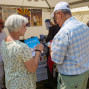 Auf dem Foto stehen vier Personen an einem Stand, der ein Buch verkauft. Eine ältere Frau und ein älterer Mann schauen gemeinsam auf ein Buch, während ein anderer Mann und ein junger Mann im Hintergrund stehen.