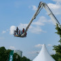 Ein Arbeitsturm mit mehreren Personen darin ist in den Himmel hineingestreckte Arm, um an einem Strommast zu arbeiten. Der Himmel ist blau mit weißen Wolken, und im Hintergrund sind Bäume und ein Gebäude zu sehen.