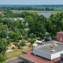 Auf dem Foto ist eine friedliche Landschaft mit einem Fluss, grünen Wäldern und einem kleinen Dorf zu sehen. Ein Park mit einer Veranstaltung und ein moderner Gebäudekomplex bilden den unteren Teil des Bildes.
