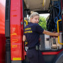 Ein Feuerwehrmann steht in einem roten Feuerwehrauto und trägt eine dunkle Uniform mit der Aufschrift "FEUERWEHR SCHWETZODER". Im Hintergrund ist die Innenausstattung des Fahrzeugs sichtbar, darunter eine weibliche Feuerwehrfrau, die sich an der Tür abstützt.