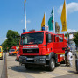 Auf einem Parkplatz vor einem belebten Gebäude steht ein roter Feuerwehrauto mit Blaulicht und der Aufschrift "FEUERWEHR". Im Hintergrund sind weitere Fahrzeuge, Flaggen und ein belebter Bereich mit Menschen zu sehen.