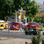 Auf dem Foto ist eine belebte Straße mit mehreren roten Feuerwehrautos und einer gelben Oldtimerkutsche zu sehen, die vor einem modernen Gebäude mit Parkplätzen und einer Fußgängerüberführung im Hintergrund steht. Grünpflanzen säumen die Straße und sorgfältig gestellte Fahrzeuge stehen zusätzlich auf dem Bürgersteig.