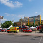 Auf dem Foto ist ein großer Veranstaltungshaushalt mit einem Feuerwehrauto und mehreren Rettungswagen vor einem belebten Bürgersteig zu sehen. Im Hintergrund ist ein blauer Himmel mit einigen Wolken und ein markantes Gebäude mit einem Turm und einigen aufgestellten Zelten zu erkennen.