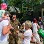 Ein Soldat lächelt und hält ein kleines Kind auf seinem Rücken, während eine Gruppe von Kindern und Erwachsenen in Militärausbildung vor einem gepanzerten Militärfahrzeug steht. Im Hintergrund sind weitere Soldaten und Kinder zu sehen, die sich zu dem Anlass versammeln.