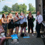 Eine Gruppe von Menschen, darunter Frauen mit roten Haaren, lacht und jubelt an einem Ufer eines Flusses. Vor ihnen steht ein Mann in einem weißen Anzug und eine große Pappaufgabe mit dem Wort "Hier" darauf.