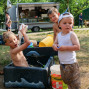 Zwei namentlich nicht erkennbare Kinder spielen ausgelassen mit Wasser in einem Planschbecken, während eine Imbisswagen im Hintergrund steht. Im Hintergrund sind weitere Menschen und ein Picknicktisch zu erkennen, was auf eine Veranstaltung im Freien hindeutet.
