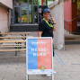 Ein Mann in einem schwarzen T-Shirt und einer Mütze lächelt in die Kamera, während er mit seiner Hand an einem Schildtipp spielt. Vor ihm steht ein Messestand mit dem Schild "INKONTAKT MESSE-BÜRO" und eine Fahrradständer.