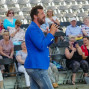 Ein Mann in einem blauen Anzug spricht in ein Mikrofon vor einer großen Menschenmenge auf einer Tribüne. Im Hintergrund sitzen viele Zuschauer, die zuhörerlich zu dem Auftritt stehen.