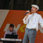 Ein Mann mit Kapuze singt mit Mikrofon auf einer Bühne vor einem orangefarbenen Hintergrund mit einem Sonnensymbol. Im Hintergrund steht ein Mann, der eine Tastatur spielt.