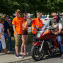 Eine Gruppe von jungen Leuten in orangefarbenen T-Shirts steht vor einer großen, schwarzen Motorrad-Tourer-Maschine, umgeben von Autos und einer älteren Dame. Auf dem Motorrad sitzt ein Mann in Leder und Stiefeln, während mehrere Personen ihre Aufmerksamkeit auf ihn richten.
