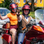 Ein Paar posiert auf einer roten Motorrad-Tour, mit einer Frau, die mit den Daumen hoch und einer Hand geben und einem Mann, der mit einem Helm und einem lächelnden Gesicht auf dem Motorrad sitzt. Im Hintergrund sind eine graue Limousine und ein Gebäude zu sehen.