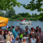 Auf dem Foto sieht man eine Menschenmenge, die am Ufer eines Flusses oder Sees steht und ein Boot beobachtet. Im Vordergrund ist ein Banner mit der Aufschrift "Öffentlich er weizen" zu sehen, während die Menschen in der Menge einige auf die Szene gerichtet sind.