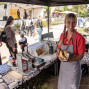 Eine lächelnde Verkäuferin in rotem Shirt und Schürze hält an einem Imbissstand ein Brötchen mit Wurst in die Kamera. Im Hintergrund findet bei sonnigem Wetter ein belebtes Fest mit Bühne, Zelten, Sitzbänken und vielen Besuchern statt.