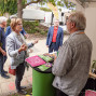 Mehrere Personen unterhalten sich an einem Informationsstand mit Broschüren und Flyern im Freien. Im Hintergrund sind ein Zelt, eine kleine Bühne, Bäume und ein Gebäude zu sehen.