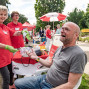 Bei einer öffentlichen Veranstaltung misst eine Frau in rotem AWO-Shirt einem sitzenden Mann den Blutdruck. Im Hintergrund sind weitere Stände, Besucher und ein rot-weißer Sonnenschirm in einer grünen Parkumgebung zu sehen.