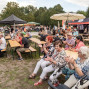 Viele Menschen sitzen bei einer Veranstaltung im Freien auf Bierbänken und schauen in Richtung einer Bühne oder Vorführung. Zwischen Zelten, Sonnenschirmen und Bäumen herrscht eine entspannte Atmosphäre, während einige Zuschauer applaudieren.